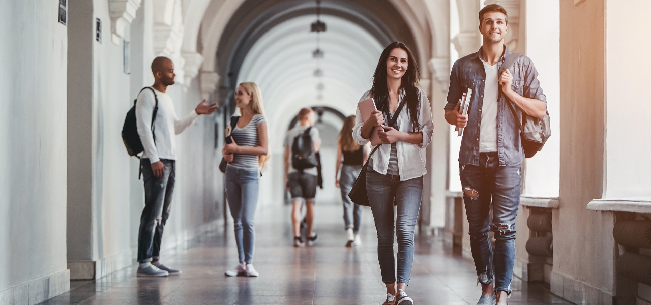 Two college students, a young man and woman, walking through a campus hallway, carrying books and backpacks.