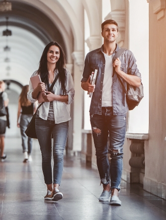 Two college students, a young man and woman, walking through a campus hallway, carrying books and backpacks.