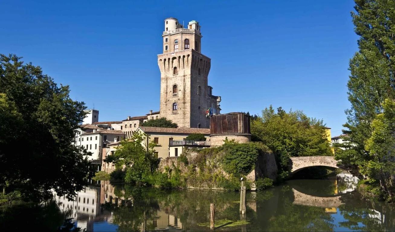 Historic stone tower with arched windows, surrounded by trees and water, reflecting the tower's image.