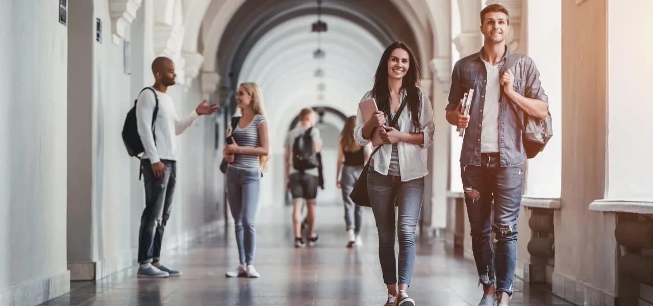 Two college students, a young man and woman, walking through a campus hallway, carrying books and backpacks.