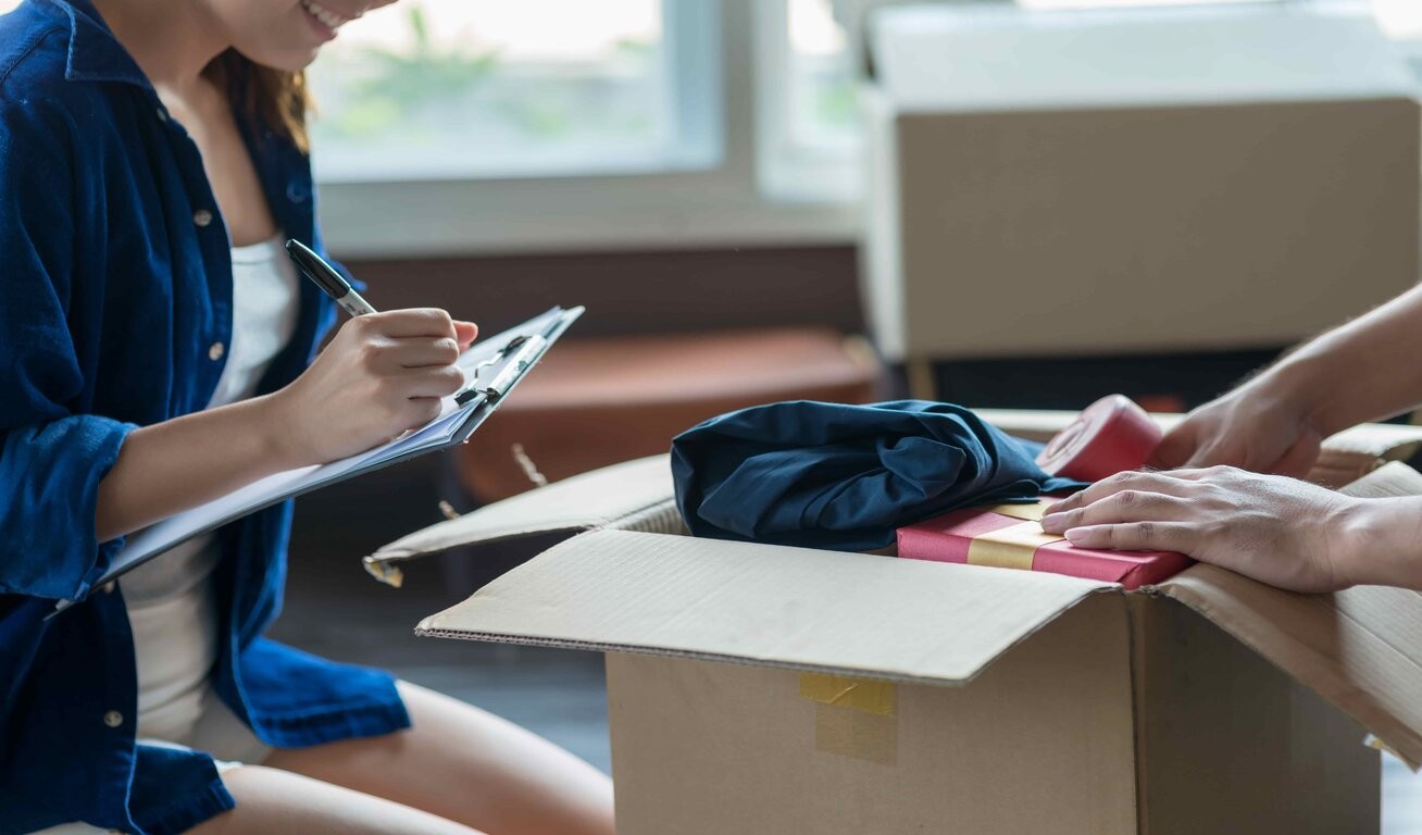A woman with a clipboard is writing notes beside a cardboard box on a table, with items like a black fabric and a red object partially visible, suggesting preparation for moving or inventory taking.