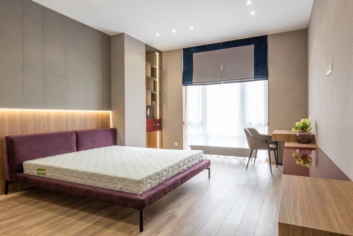 A bare mattress on an upholstered bed frame in a bright, empty bedroom