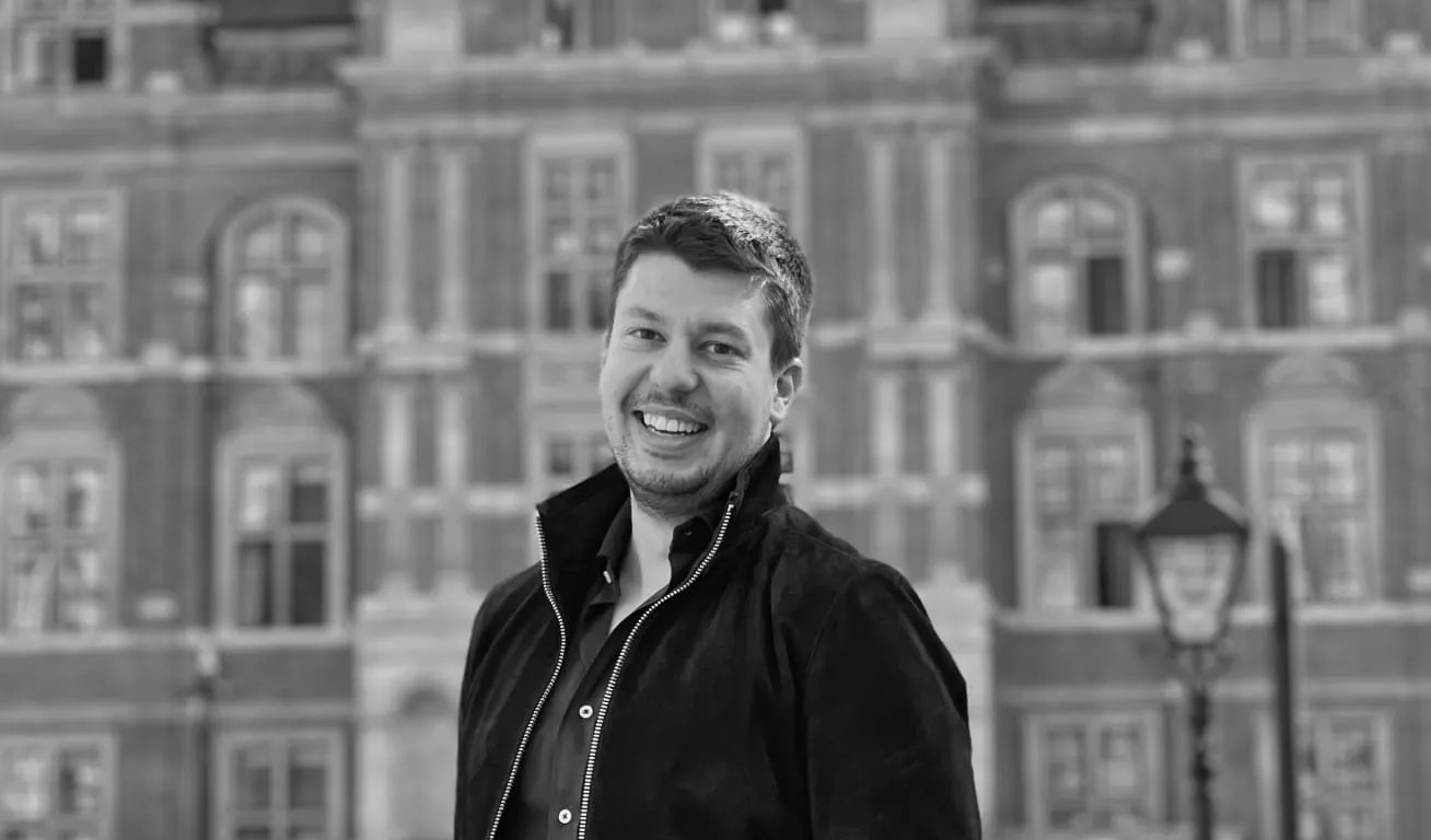 Portrait of a Happy Man Outdoors with Historical Building Behind Smiling man in casual clothing standing in front of a brick building with classic architecture and a street lamp visible in the background.